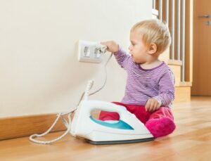 Kuvassa on pieni lapsi tutkimassa silitysrautaa ja sen johtoa lähellä seinässä olevaa pistorasiaa, mikä korostaa lapsiturvallisuuden tärkeyttä kotona ja vaaroja, joita sähkölaitteet voivat lapsille aiheuttaa.